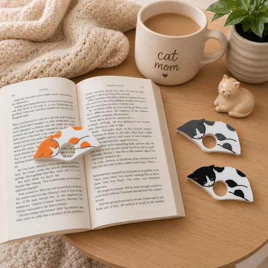 Two white spotted cats in black and gray are displayed on a wooden table next to an open book with the orange and white spotted cat, a ceramic mug with 'cat mom' printed on it, and a small cat figurine.