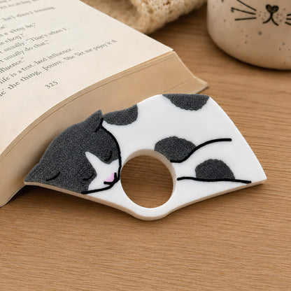 A close up of the sleeping gray and white cat page holder, resting on a wooden tabletop next to an open book.