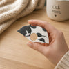 A hand holds a small cat-shaped reading ring accessory with a gray and white sleeping cat illustration, set on a light wood table with a beige knit blanket and a mug in the background.