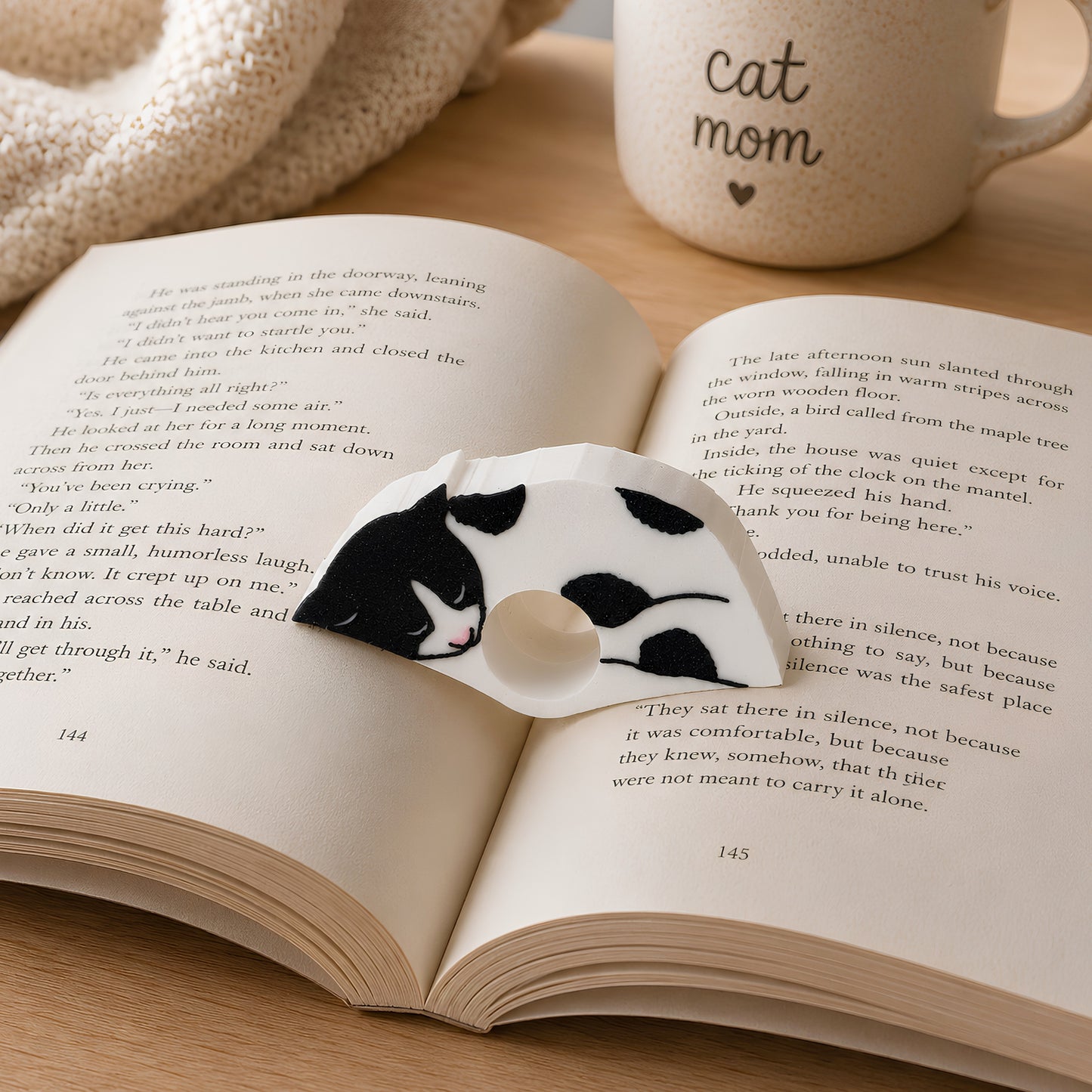 An open book spread on a light surface, featuring a black and white cat sleeping on the pages. A white mug in the background has the words 'cat mom' printed on it.
