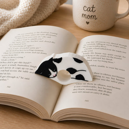An open book spread on a light surface, featuring a black and white cat sleeping on the pages. A white mug in the background has the words 'cat mom' printed on it.