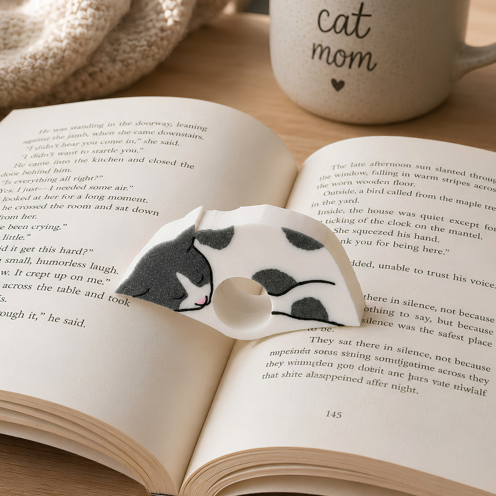 An open book spread on a light surface, featuring a gray and white cat sleeping on the pages. A white mug in the background has the words 'cat mom' printed on it.