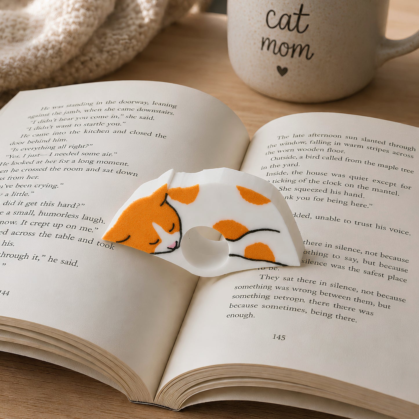 An open book spread on a light surface, featuring a orange and white cat sleeping on the pages. A white mug in the background has the words 'cat mom' printed on it.