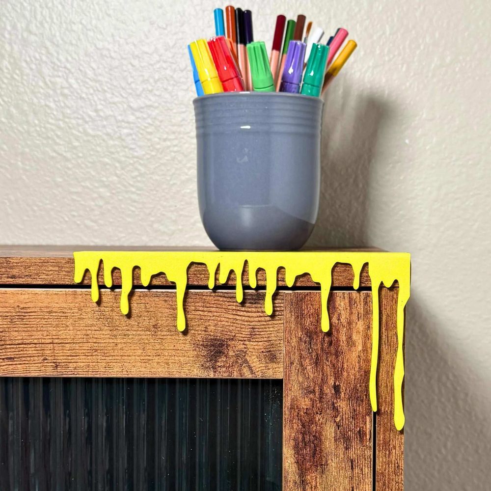 Bright yellow paint dripping art decor on the edge of a cabinet next to a mug full of colorful markers and pencils