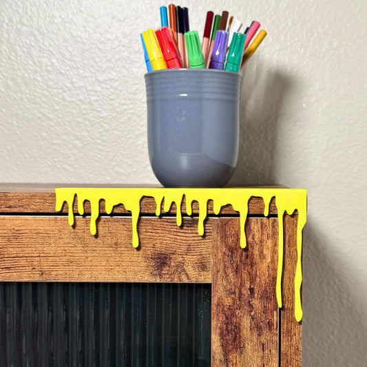 Bright yellow paint dripping art decor on the edge of a cabinet next to a mug full of colorful markers and pencils