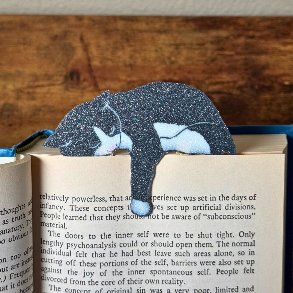 Gray and white tuxedo cat bookmark shown in a book holding onto a page with its paw dangling