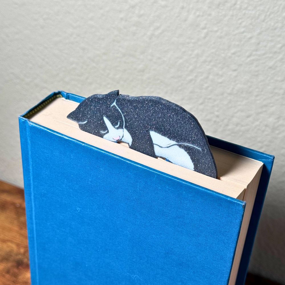 Grey tuxedo cat bookmark shown in a closed book taking a nap on top