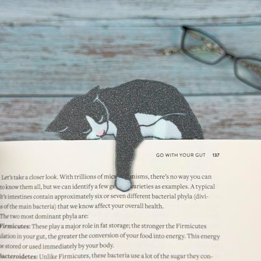 Gray and white tuxedo cat bookmark shown in a book holding onto a page with its paw dangling
