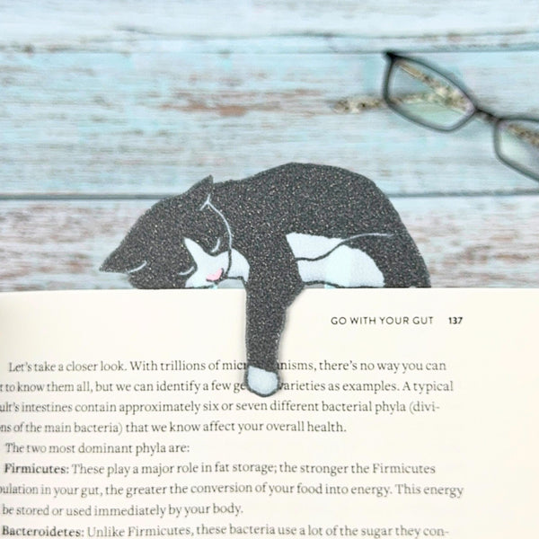 Gray and white tuxedo cat bookmark draped over a page, sleeping with one paw dangling down the book
