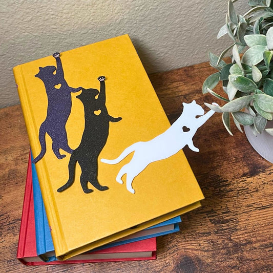 Fun cat bookmarks with a hanging paw and pink toe beans. Bookmarks shown in purple and black sparkle, as well as white