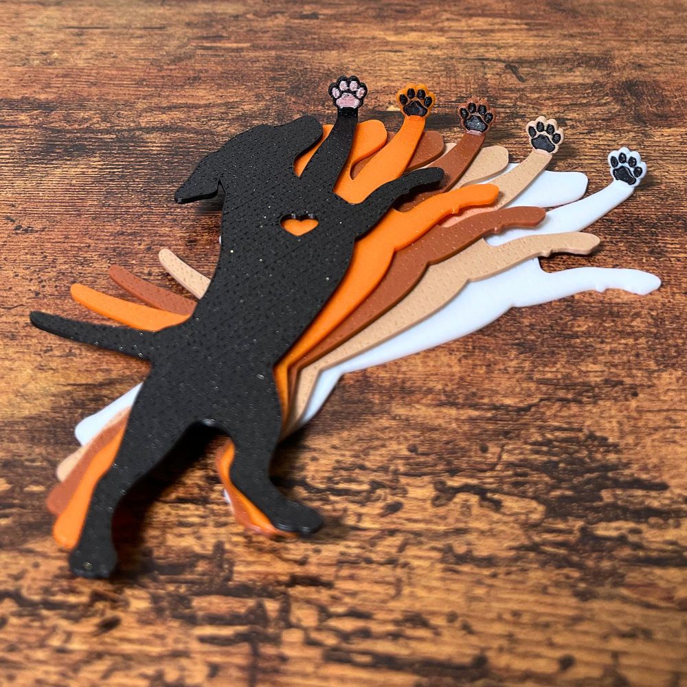 Five dog bookmarks in orange, white, tan, brown, and black sparkle laying down, showing their adorable paw toe beans on a wooden surface