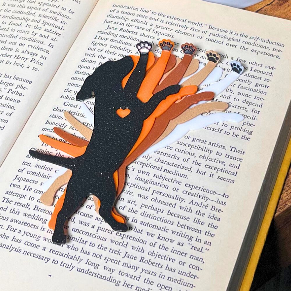 Five dog bookmarks in orange, white, tan, brown, and black sparkle laying on top of an open book, showing their adorable paw toe beans