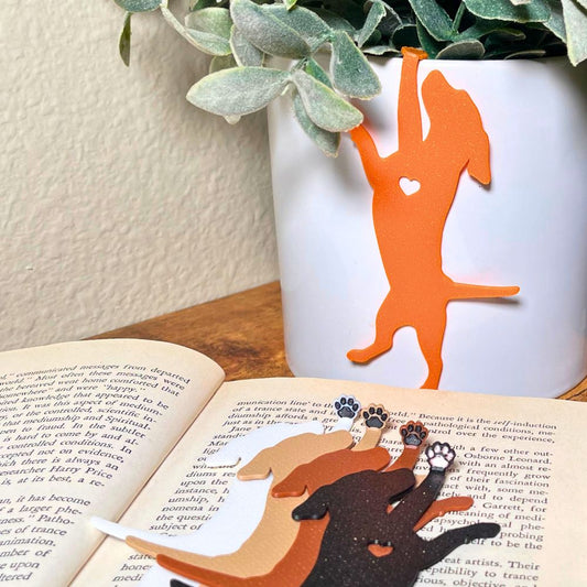 Orange hanging dog bookmark dangling from a planter, with four additional dog bookmarks in white, tan, brown, and black sparkle laying on an open book, showing their adorable paw toe beans