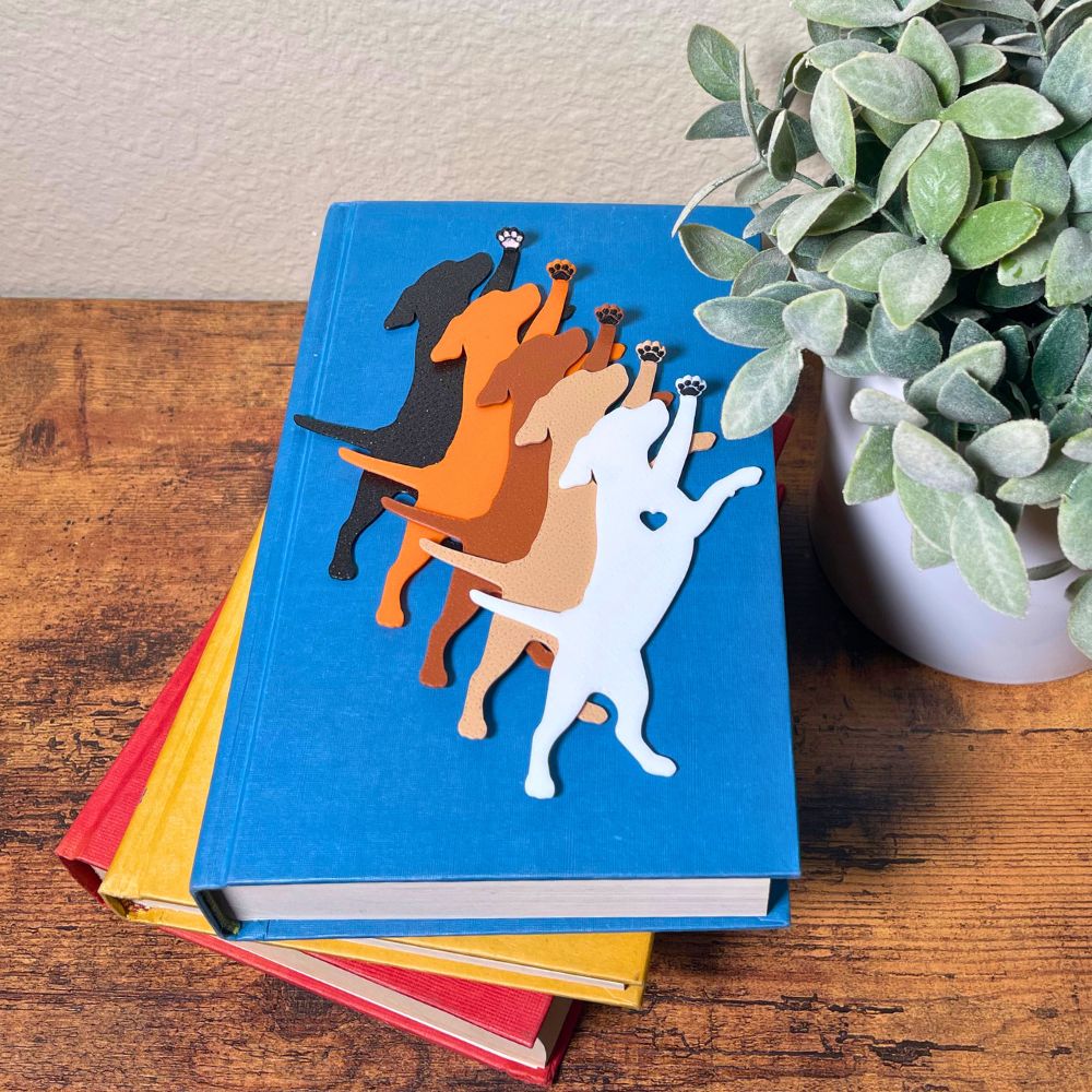 Five dog bookmarks in orange, white, tan, brown, and black sparkle laying on top of a book, showing their adorable paw toe beans