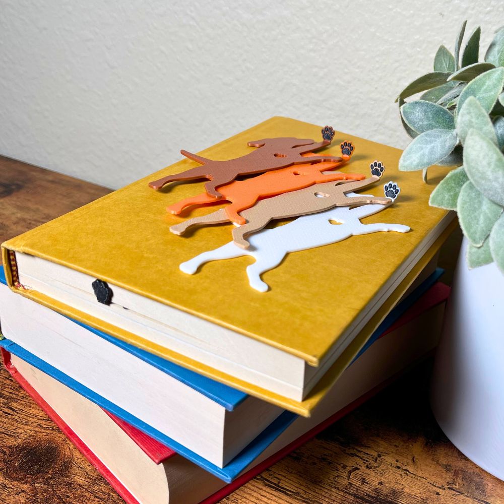 Black sparkle hanging dog bookmark inside the book only showing the top of the paw sticking out, with four additional dog bookmarks in white, tan, brown, and orange laying on a book, showing their adorable paw toe beans