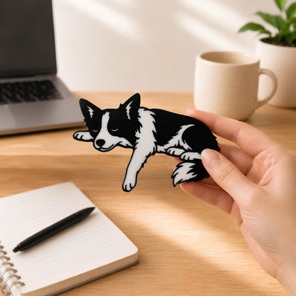 Hand holding the small Border Collie shelf pet at a sunlit desk with a laptop, mug, and open notebook