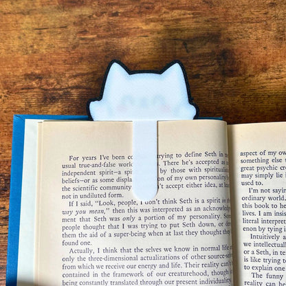 The backside view of the kawaii style cat bookmark clipped onto the page of a blue book from an angle