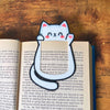 Cute kawaii style cat bookmark waving hello clipped onto the page of a blue book showing off its cute pink toe beans