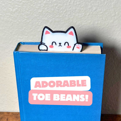 Cute kawaii style cat bookmark waving hello from a blue book showing off its cute pink toe beans