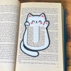 Cute kawaii style cat bookmark waving hello from inside a blue book showing off its cute pink toe beans