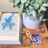 Four colors of fun monkey bookmarks hanging from a book and on a shelf
