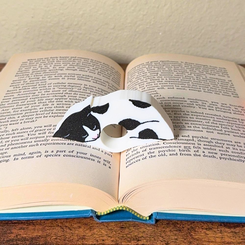 Adorable sleeping black and white spotted cat book page holder shown in a book