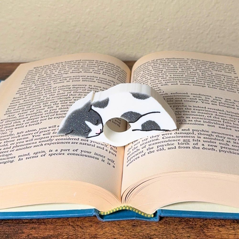 Adorable sleeping gray and white spotted cat book page holder shown in a book