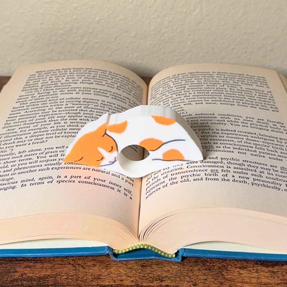 Adorable sleeping orange and white spotted cat book page holder shown in a book