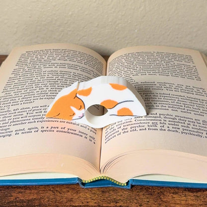 Adorable sleeping orange and white spotted cat book page holder shown in a book