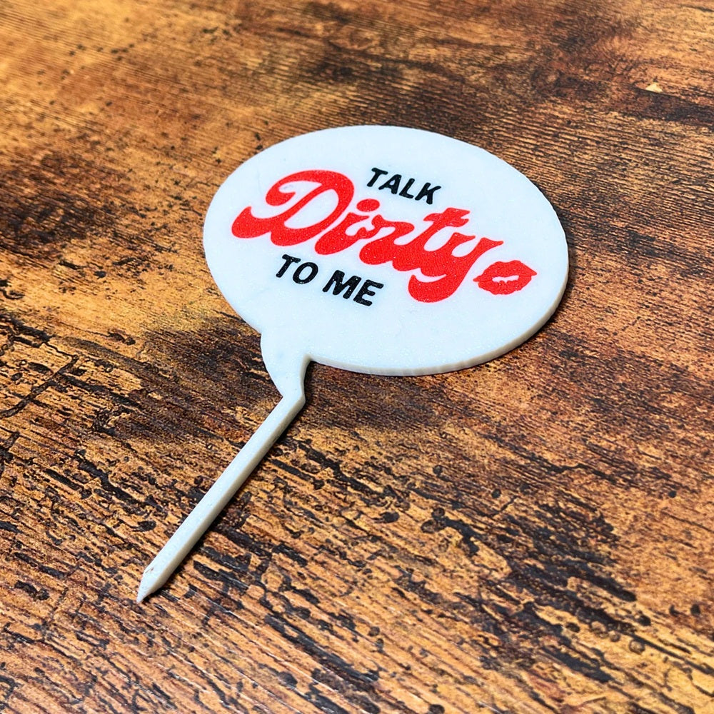 The punny Talk Dirty to Me plant stake on a wooden surface