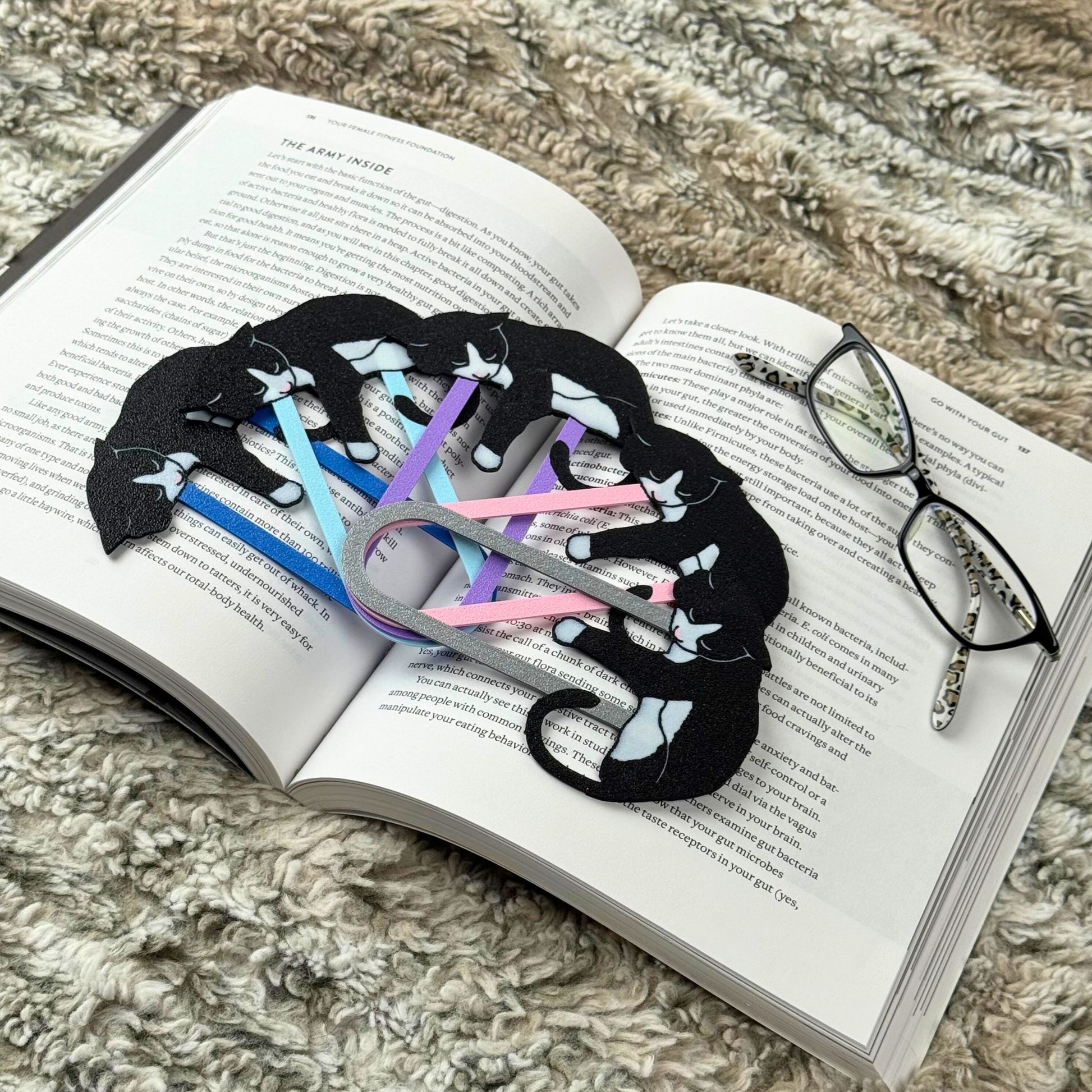 Set of tuxedo cat bookmarks in 5 different clip colors on an open book with glasses beside it.