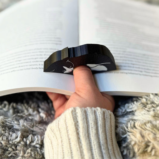 Hand holding a book open with a black and white cat patterned thumb book holder ring.