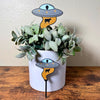 2 different size UFO Plant stakes with alien inside using it's golden beam to pull up a cow stuck inside a house plant and one in front of the house plant