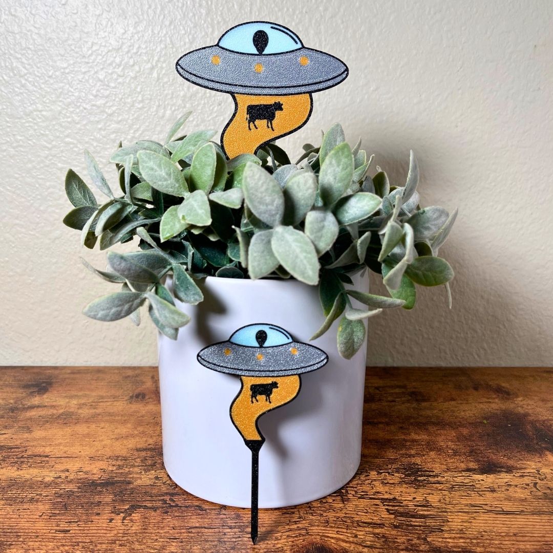 2 different size UFO Plant stakes with alien inside using it's golden beam to pull up a cow stuck inside a house plant and one in front of the house plant